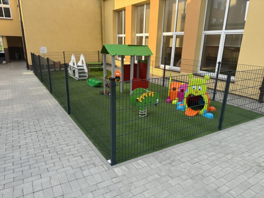 Bezpieczny plac zabaw dla dzieci, z kolorowymi zjeżdżalniami, huśtawkami i zabawkami na sztucznej trawie, otoczony ogrodzeniem, znajdujący się w przestrzeni edukacyjnej.