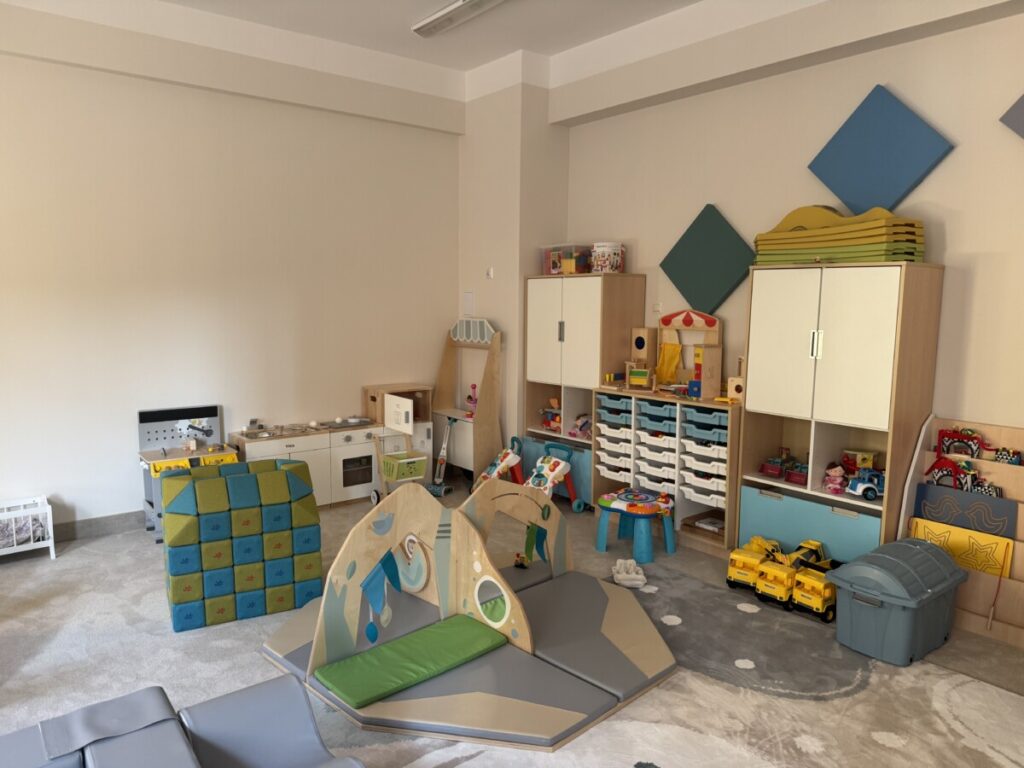 Przestronny pokój zabaw dla dzieci z różnorodnymi zabawkami, w tym kuchnią, klockami i mebelkami do przechowywania. W centrum znajduje się kolorowa konstrukcja do wspinania oraz poduszki do zabawy na miękkiej podłodze. Jasne ściany i atrakcyjne dekoracje tworzą przyjazną atmosferę dla maluchów.