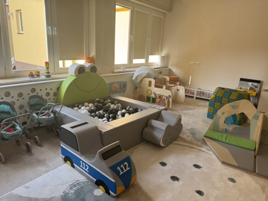 Pokój zabaw dla dzieci z kolorowymi zabawkami, strefą do zabawy w kulkach oraz meblami w kształcie samochodów i chmur. W tle wózki dla lalek i drewniane elementy edukacyjne. Idealne miejsce do kreatywnej zabawy i rozwoju maluchów.