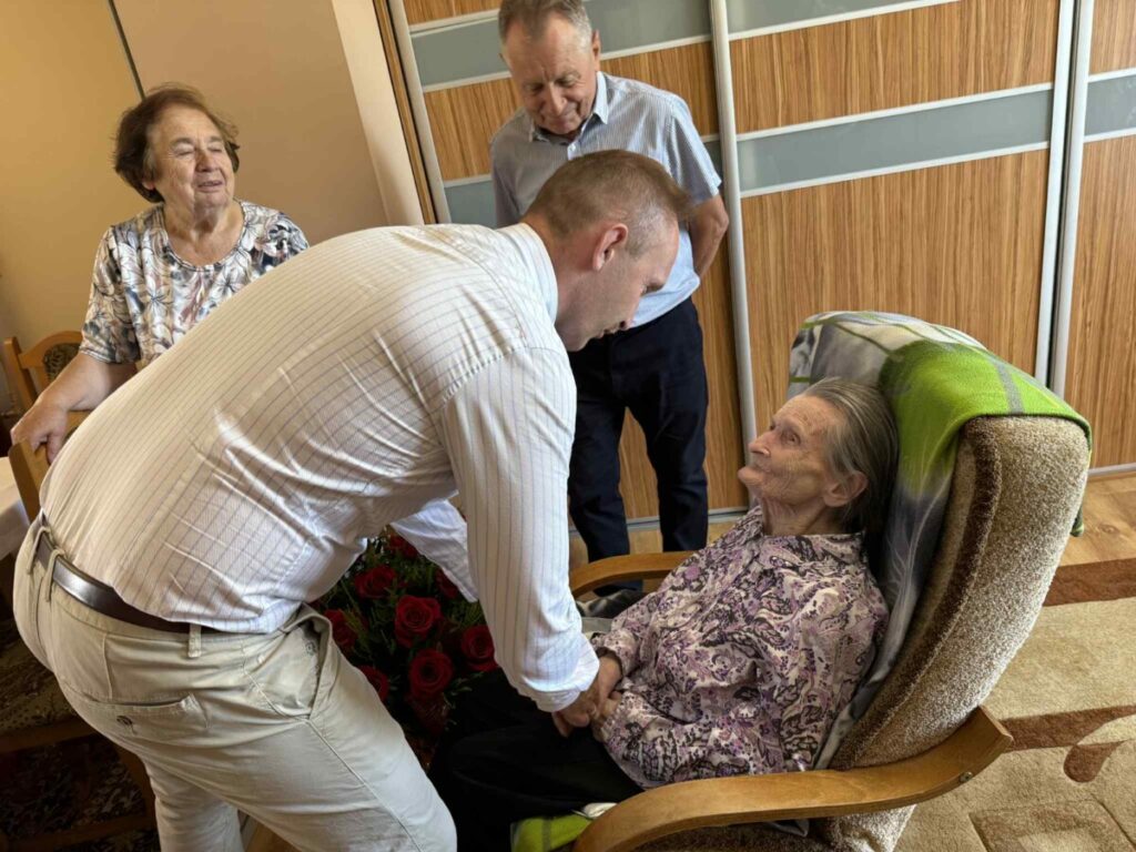 Osoba starsza siedzi w fotelu, otrzymując wizytę od sympatycznego mężczyzny, który rozmawia z nią, trzymając jej dłonie. W tle widoczni są inni członkowie rodziny, w tym kobieta i mężczyzna, tworząc atmosferę wsparcia i bliskości. W pomieszczeniu znajdują się kwiaty, co sugeruje okazję do świętowania.