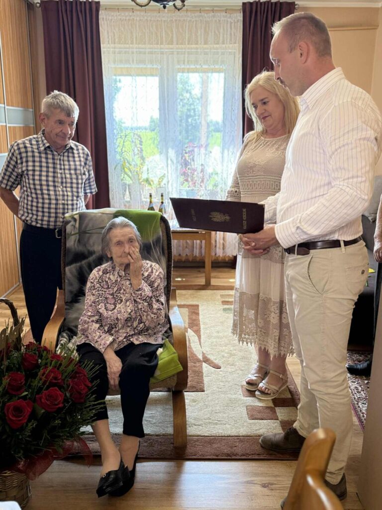 Rodzina w mieszkaniu, gdzie starsza kobieta siedzi na fotelu z bukietem czerwonych róż, podczas gdy trzech krewnych, w tym dwóch mężczyzn i jedna kobieta, z zainteresowaniem ogląda dokumenty. W tle widać okno z roślinami i domowe wnętrze.