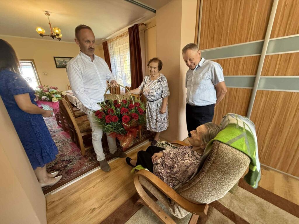 Rodzina odwiedza starszą kobietę, przynosząc jej bukiety kwiatów i wspólnie spędzając czas w przytulnym wnętrzu domu.