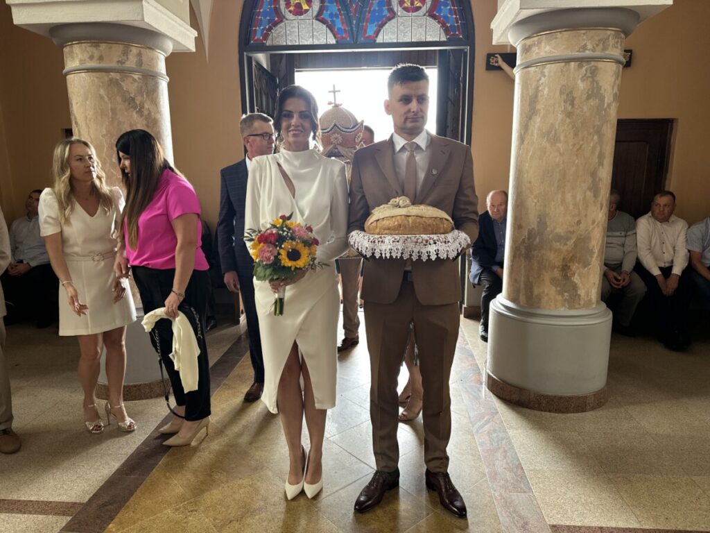 Młoda para w kościele, szczęśliwie wchodząca z kwiatami w rękach. Kobieta w eleganckiej, białej sukni trzyma bukiet z kolorowych kwiatów, a mężczyzna w brązowym garniturze niesie chleb na talerzu, ozdobionym koronką. W tle widać gości ślubnych siedzących w ławkach.