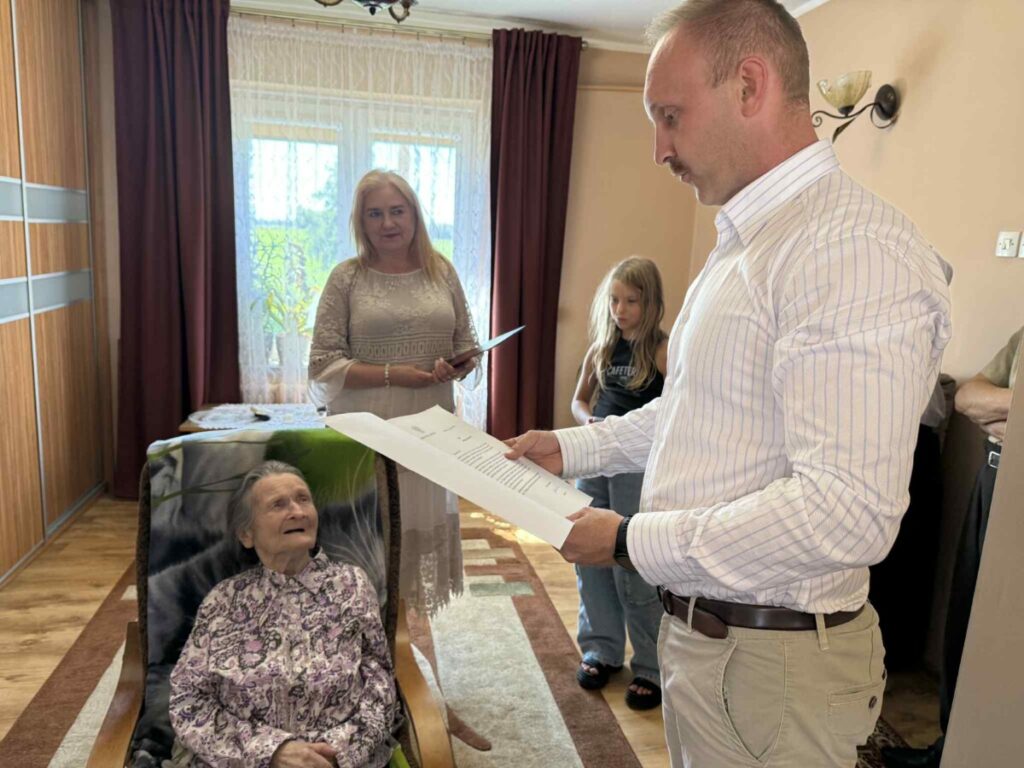 Spotkanie rodzinne, podczas którego mężczyzna czyta dokument przed starszą kobietą siedzącą w fotelu. Obok stoi kobieta z kartką, a w tle błąka się dziecko. Wnętrze jasne, z drewnianymi elementami i zasłonami.