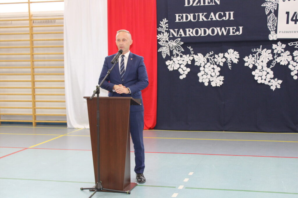 Mężczyzna w garniturze przemawia z mównicy podczas uroczystości Dnia Edukacji Narodowej w sali sportowej, z dekoracjami w kolorach polskiej flagi w tle.