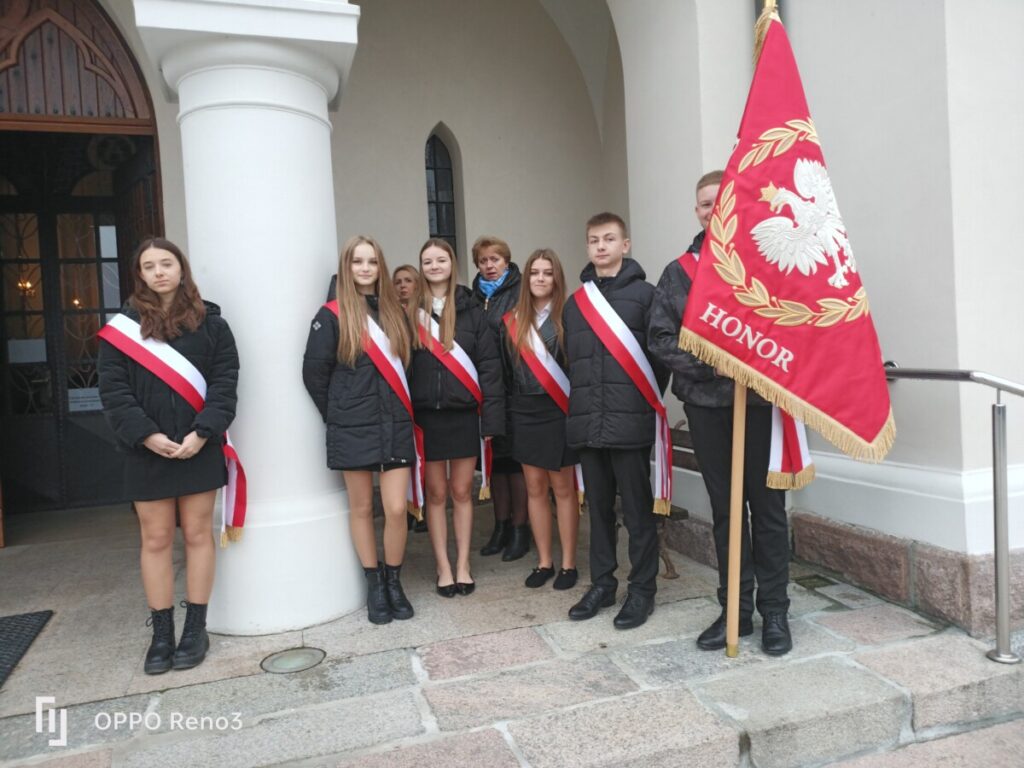 Grupa młodzieży w galowych strojach stoi przed wejściem do budynku, trzymając flagę z orłem białym i inskrypcją "HONOR". Wszyscy mają na sobie czerwono-białe wstęgi, symbolizujące patriotyzm.