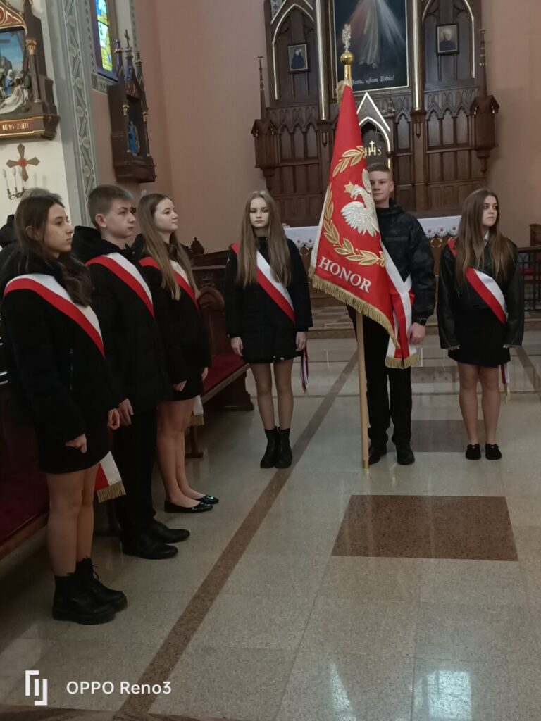 Grupa młodzieży w mundurach, trzymająca sztandar w kościele, podczas ceremonii. Uczniowie w odzieży formalnej z biało-czerwonymi ozdobami, stoją w rzędzie obok sztandaru, na tle drewnianego ołtarza.