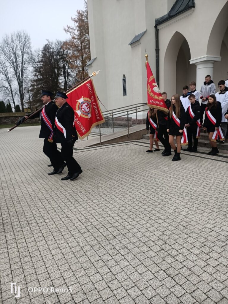 Uczestnicy ceremonii marszowej przed kościołem, niosący sztandary, w tym flagi z napisami "Bóg na Cześć" i "Honor". Grupa młodzieży w strojach galowych z biało-czerwonymi wstęgami, widoczna w tle, w tle drzewa i architektura budynku.
