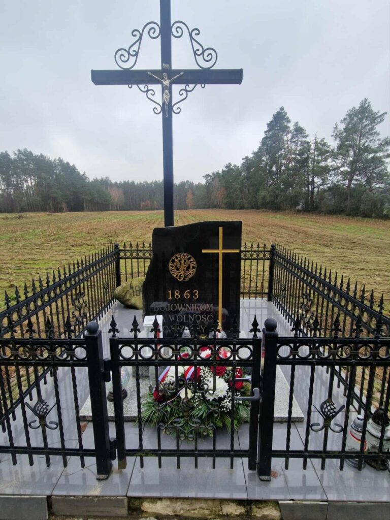 Pomnik w hołdzie powstańcom z 1863 roku, znajdujący się na tle lasu. Krzyż metalowy z figurą Jezusa, otoczony eleganckim ogrodzeniem, z kwiatami i zniczami na płycie nagrobnej. Idealne miejsce pamięci i refleksji.
