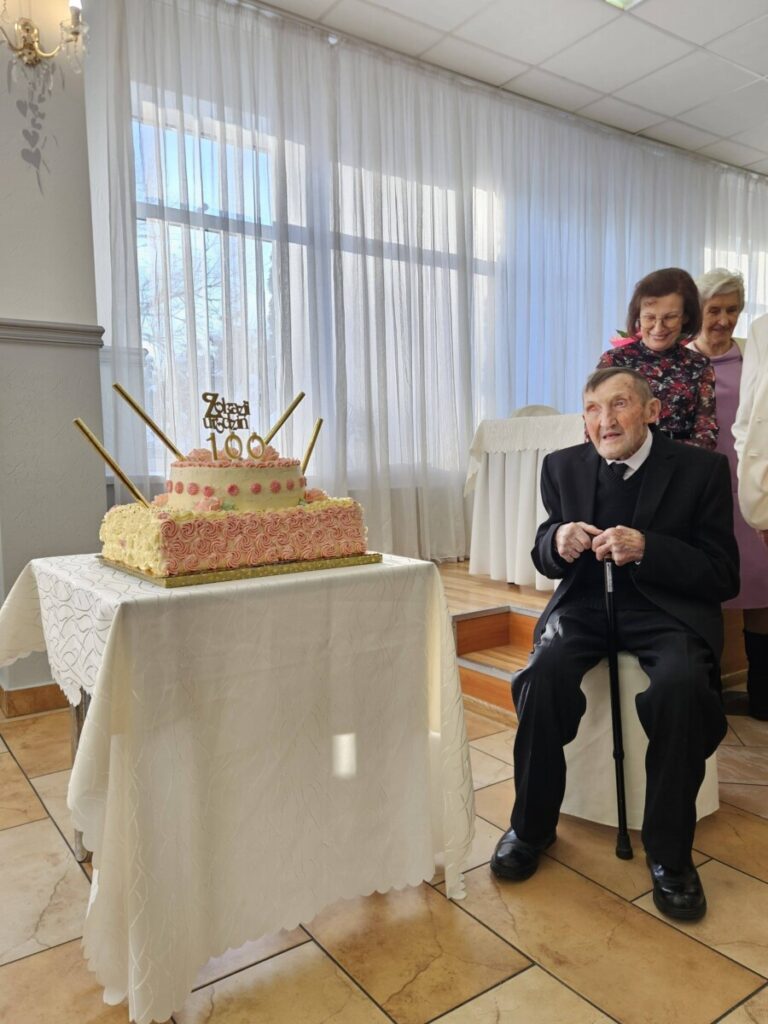 Mężczyzna siedzący przy stole z tortem na swoje setne urodziny, otoczony przez trzy kobiety w tle. Tort ozdobiony różowymi różami i świeczkami, z napisem "100". Jasne, przestrzenne wnętrze z zasłonami.